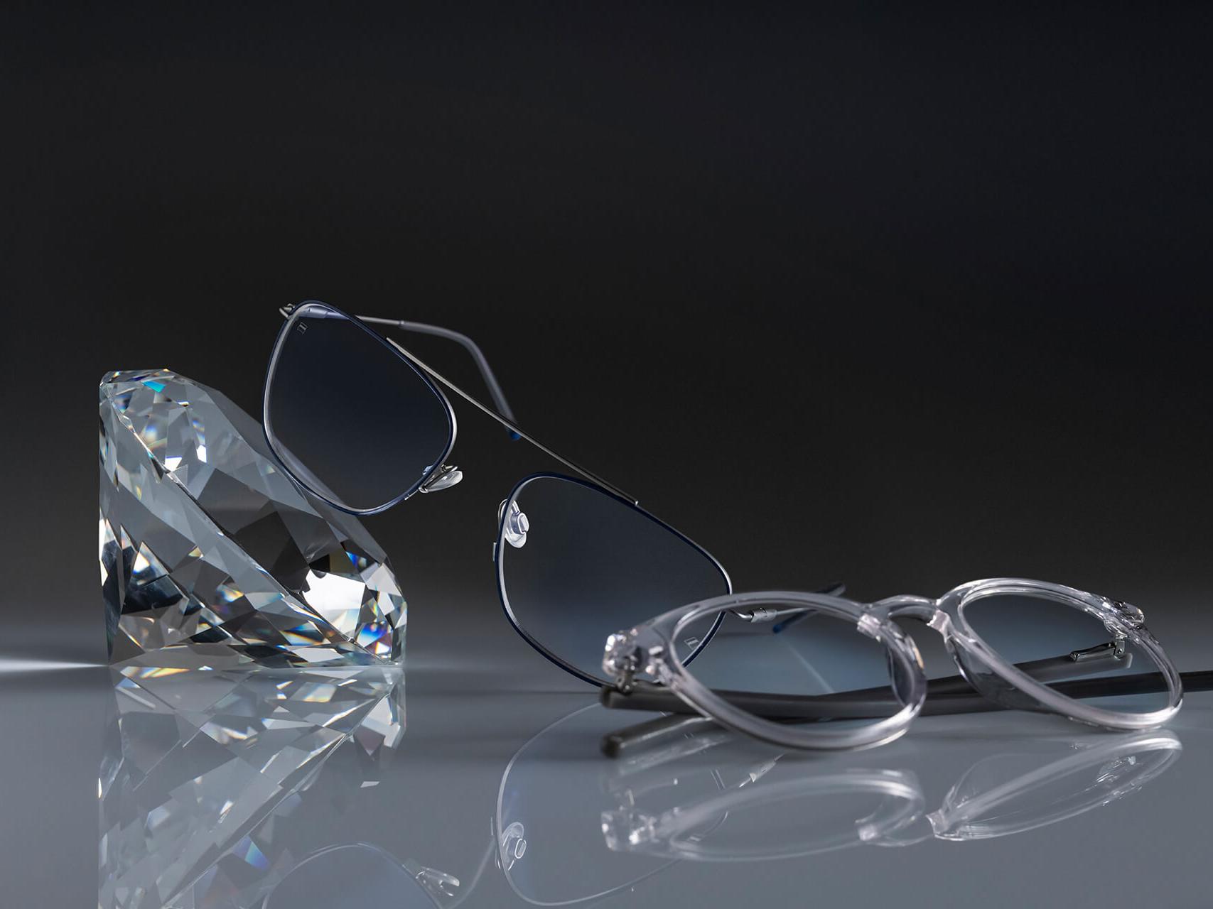 Dva para naočala sa ZEISS lećama koje imaju najtvrđi ZEISS zaštitni sloj svih vremena – DuraVision® Platinum. Jedan par naočala naslonjen je na kristal, a drugi leži ravno na tlu. Oba imaju prozirne leće bez plavkastog odsjaja.
