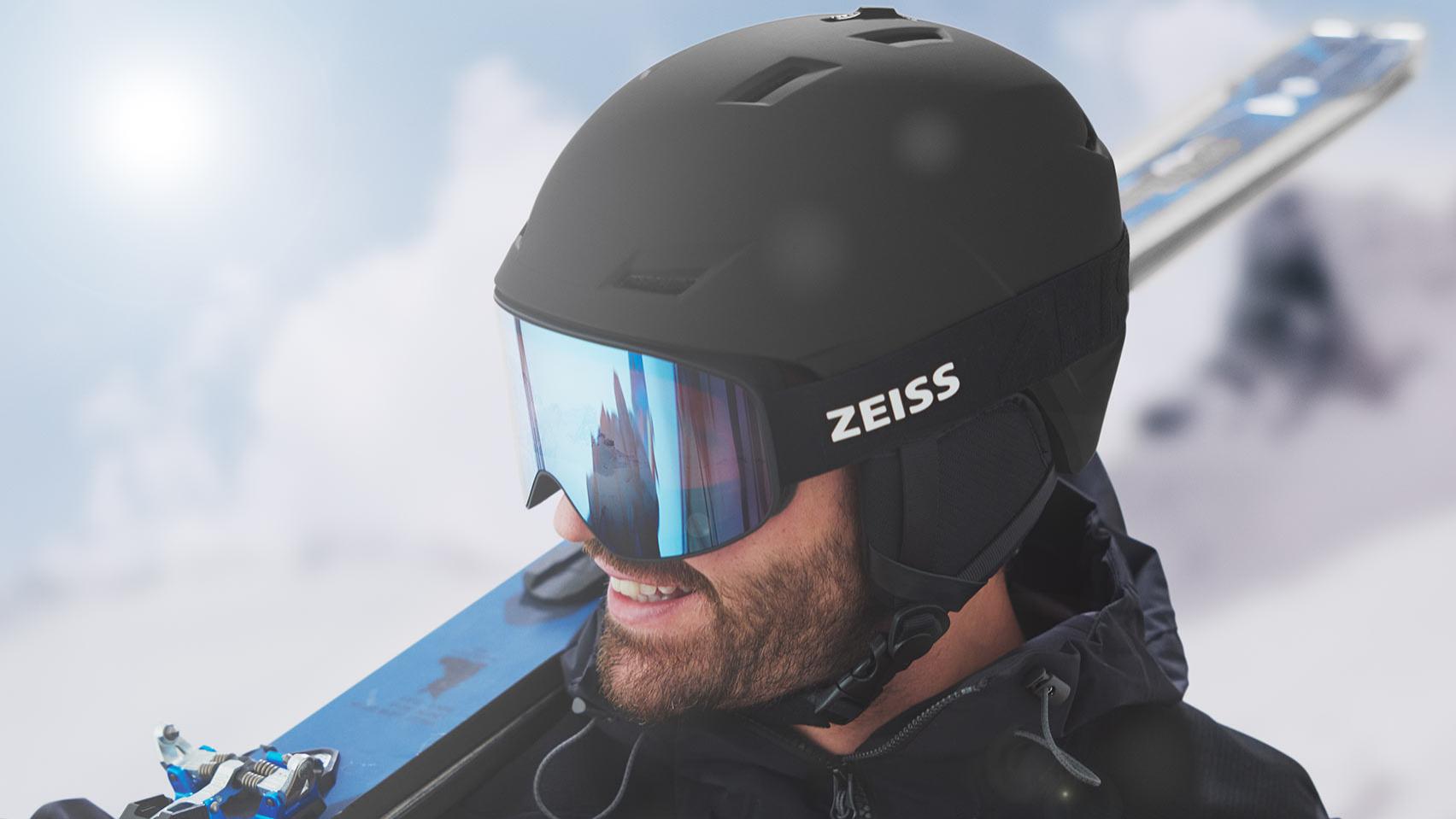 Muškarac nosi crnu kacigu s plavim reflektirajućim ZEISS naočalama za skijanje, ima skije preko desnog ramena.